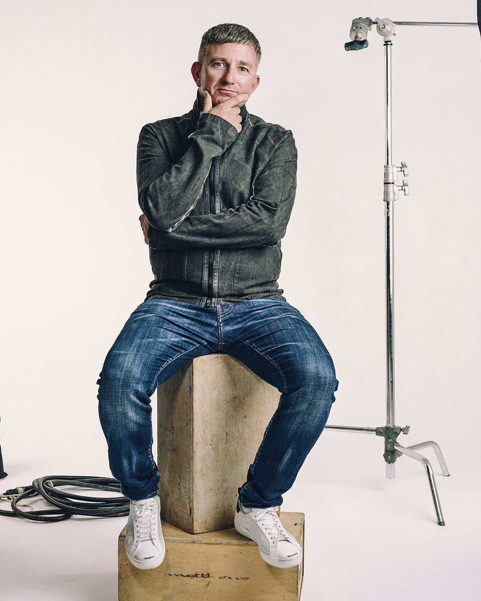 Matthew Benjamin seated on a wooden box in a photo studio, hand resting at his chin, a light stand beside him